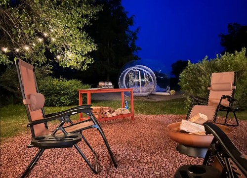 A cozy outdoor space at night with chairs, a fire pit, and a transparent bubble tent surrounded by greenery.