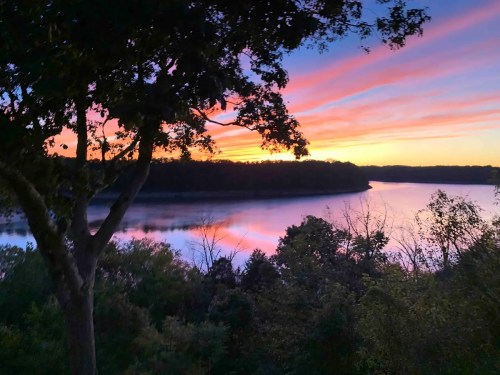 A serene sunset over a calm river, with vibrant pink and orange skies reflecting on the water, framed by trees.