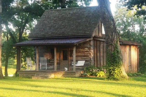 A rustic wooden cabin with a porch, surrounded by greenery and trees, featuring rocking chairs and a small white cat.
