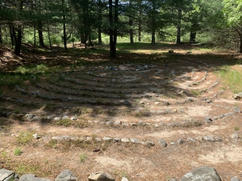 A stone labyrinth set in a forest, surrounded by trees and grass, with winding paths marked by rocks.