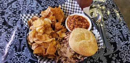 A plate of pulled pork on a bun, served with potato chips and baked beans, on a patterned tablecloth.