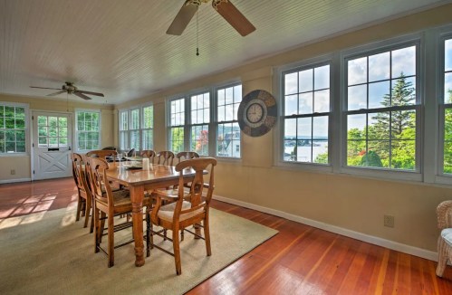 Bright dining area with a wooden table and chairs, large windows, and a view of greenery and water outside.