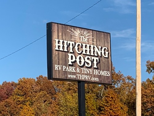 Sign for "The Hitching Post" RV Park & Tiny Homes, featuring a rustic design against a clear blue sky.