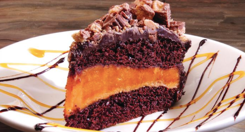 A slice of chocolate cake with a rich orange filling, topped with chocolate and crushed candy pieces on a decorative plate.