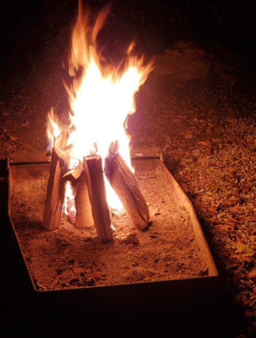 A bright campfire with flames rising from logs in a sandy fire pit, surrounded by dark outdoor surroundings.