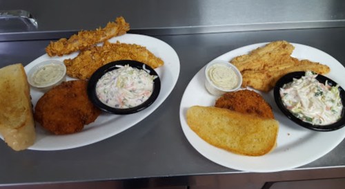 Two plates of fried fish with coleslaw, hushpuppies, and dipping sauce, served on a stainless steel countertop.