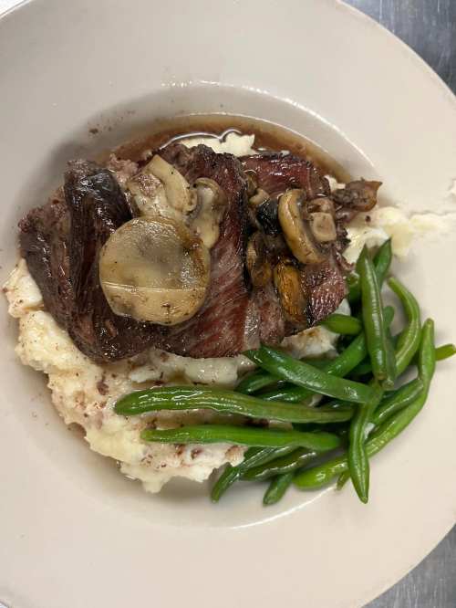 A plate of mashed potatoes topped with steak and mushrooms, accompanied by green beans.