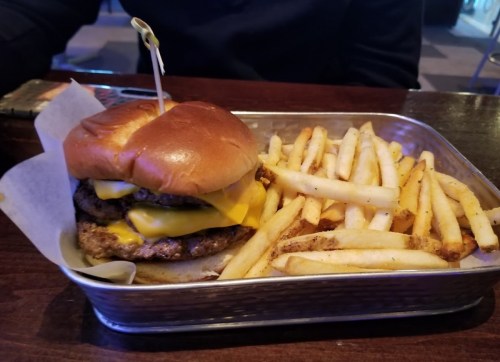 A cheeseburger with two patties and melted cheese, served with a side of golden French fries on a metal tray.