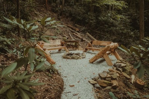 A serene forest clearing with a stone fire pit surrounded by wooden benches and gravel path.