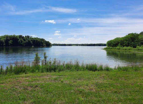A serene lake surrounded by lush greenery under a clear blue sky with scattered clouds.