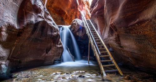 A wooden ladder leads to a waterfall in a narrow canyon, surrounded by red rock formations and flowing water.