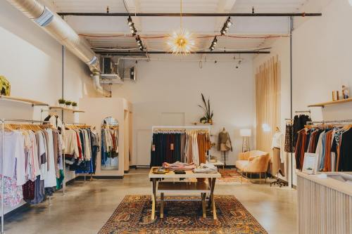 A stylish boutique interior featuring clothing racks, a central table, and cozy seating, with warm lighting and decor.