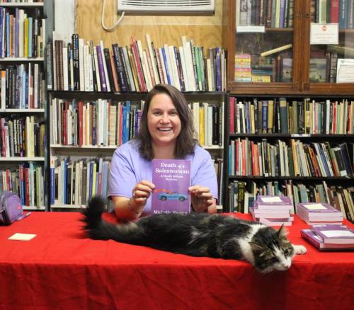 A smiling woman holds a book at a table with a cat lounging beside her, surrounded by shelves of books.