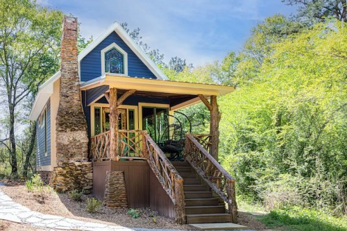 A charming blue cottage with a stone chimney, surrounded by greenery and featuring a welcoming porch and steps.
