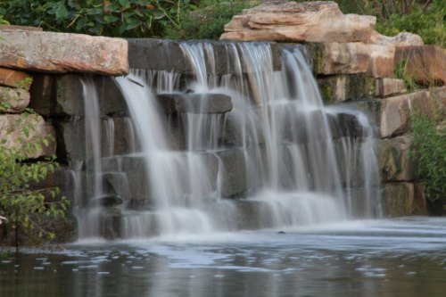 A serene waterfall cascading over stone steps into a calm pool, surrounded by lush greenery.