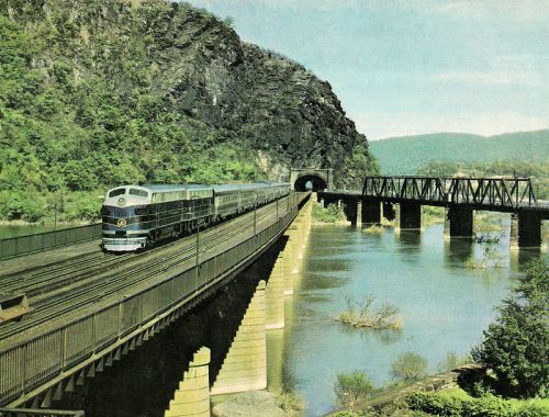 A vintage train travels along a bridge by a river, with a rocky landscape and a tunnel in the background.