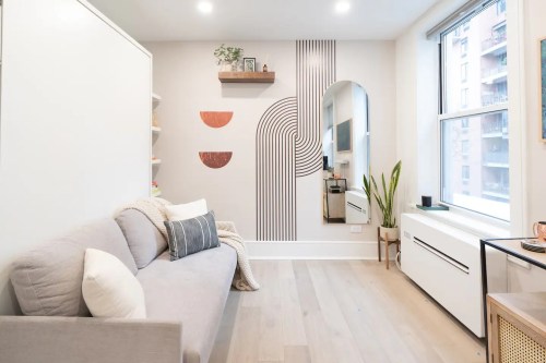 Cozy living space with a gray sofa, decorative wall art, a mirror, and a plant by the window. Bright and modern design.