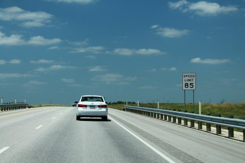 A white car drives on a highway with a sign indicating a speed limit of 85 mph under a blue sky with clouds.