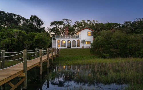 A charming house with a deck, surrounded by greenery, beside a calm waterway at dusk.