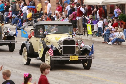 A vintage car in a parade, surrounded by spectators and colorful flags, with children waving and enjoying the event.
