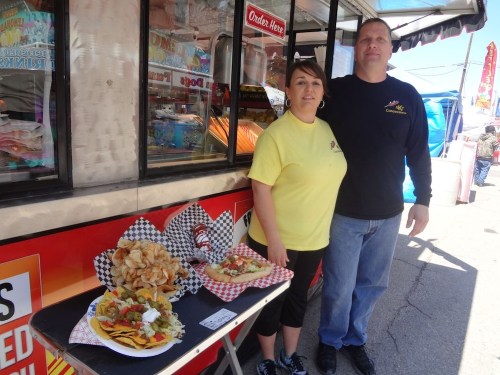 A man and woman stand in front of a food truck, showcasing a variety of colorful dishes on a table.
