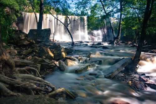 A serene waterfall cascades over rocks, surrounded by lush greenery and flowing water in a tranquil natural setting.