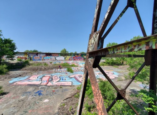 A view of a graffiti-covered area with greenery, framed by a rusted metal structure under a clear blue sky.