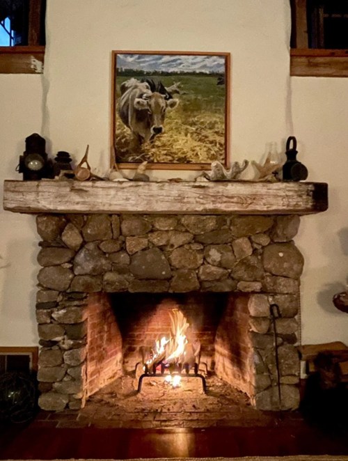 A cozy stone fireplace with a crackling fire, framed by a cow painting above the mantel. Warm and inviting atmosphere.