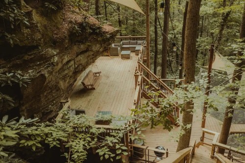 A wooden deck surrounded by trees, featuring seating areas and a rock wall in a serene forest setting.