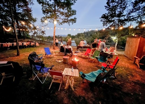 A cozy outdoor gathering with chairs around a fire pit, string lights overhead, and people socializing in the evening.