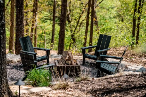 A cozy outdoor fire pit area with four black chairs surrounded by trees and greenery.