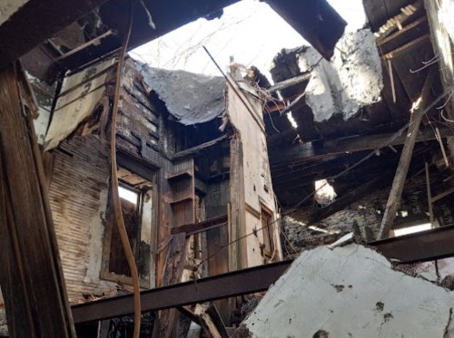 Interior of a dilapidated building with collapsed walls and roof, showing exposed beams and debris.