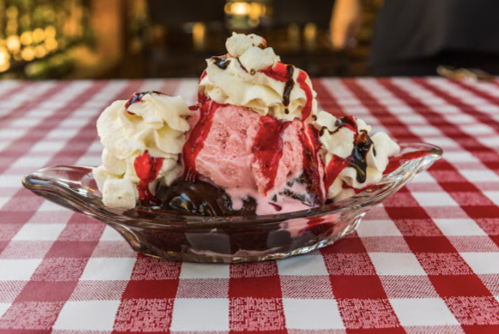 A glass dish filled with pink ice cream, whipped cream, chocolate syrup, and cherries, on a red and white checkered tablecloth.