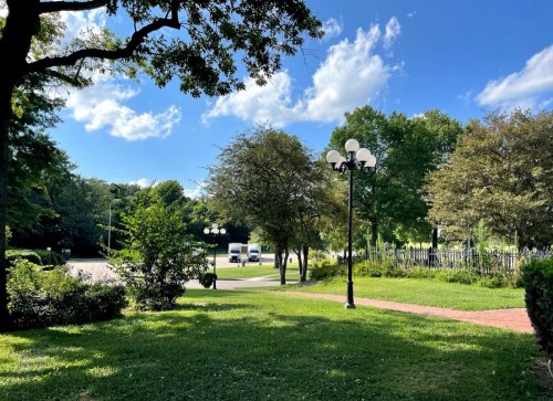 A sunny park scene with green grass, trees, and decorative street lamps under a blue sky with fluffy clouds.