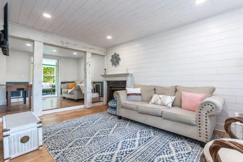 Cozy living room with a beige sofa, blue patterned rug, and a fireplace, featuring white shiplap walls and wooden beams.
