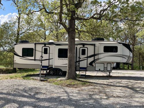 A white RV parked under a tree in a gravel area, surrounded by greenery and blue skies.