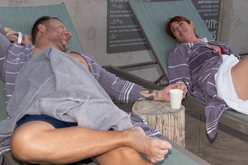 A man and woman relax on lounge chairs, smiling and enjoying drinks, wrapped in towels at a spa-like setting.
