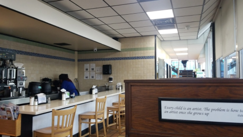 Interior of a cafe with empty seating, a coffee station, and a quote on the wall about childhood and artistry.