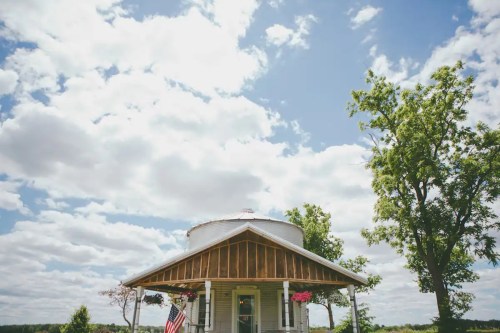 A charming round house with a porch, surrounded by trees and under a bright blue sky with fluffy clouds.