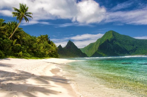 A serene beach with white sand, lush greenery, and mountains under a blue sky with fluffy clouds.