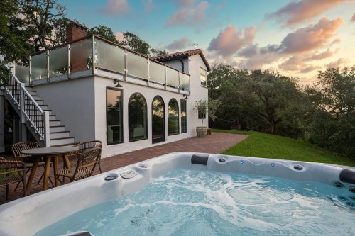 A hot tub in the foreground with a modern house and patio in the background, surrounded by greenery and a colorful sky.