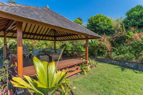 A wooden gazebo with a thatched roof surrounded by lush greenery and colorful plants on a sunny day.