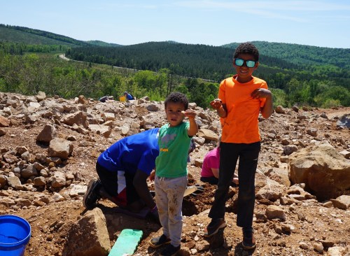 Two children stand on a rocky hillside, one holding a small object, with green hills and a road in the background.