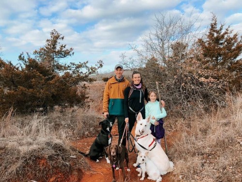 A man, woman, and girl stand on a trail with three dogs, surrounded by trees and a cloudy sky.