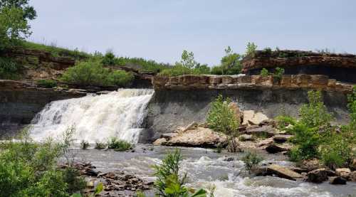 A waterfall cascading over rocky cliffs, surrounded by lush greenery and a flowing river under a clear sky.