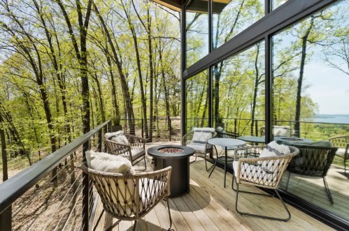A modern outdoor deck with cozy seating and a fire pit, surrounded by lush green trees and large glass windows.