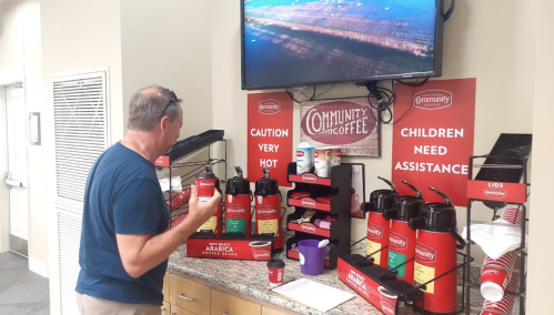 A person stands at a coffee station with various coffee dispensers and a TV screen displaying a landscape.