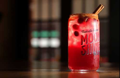 A vibrant red cocktail in a glass with a cinnamon stick, garnished with cherries, set against a blurred background.