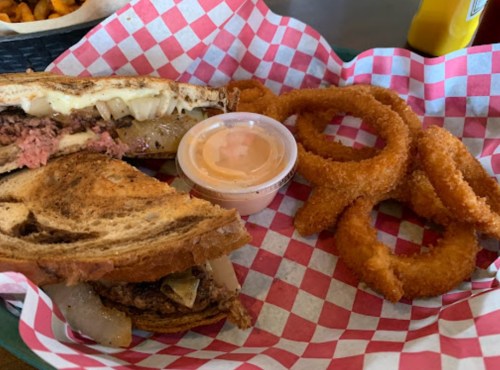 A sandwich with meat and onions on toasted bread, served with onion rings and a small cup of dipping sauce.