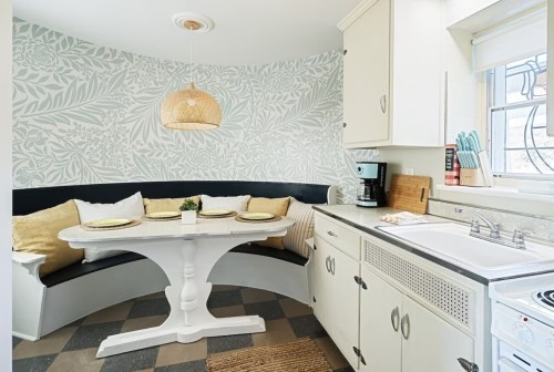 Cozy kitchen nook with a round table, cushioned seating, and floral wallpaper, featuring a pendant light and kitchen appliances.